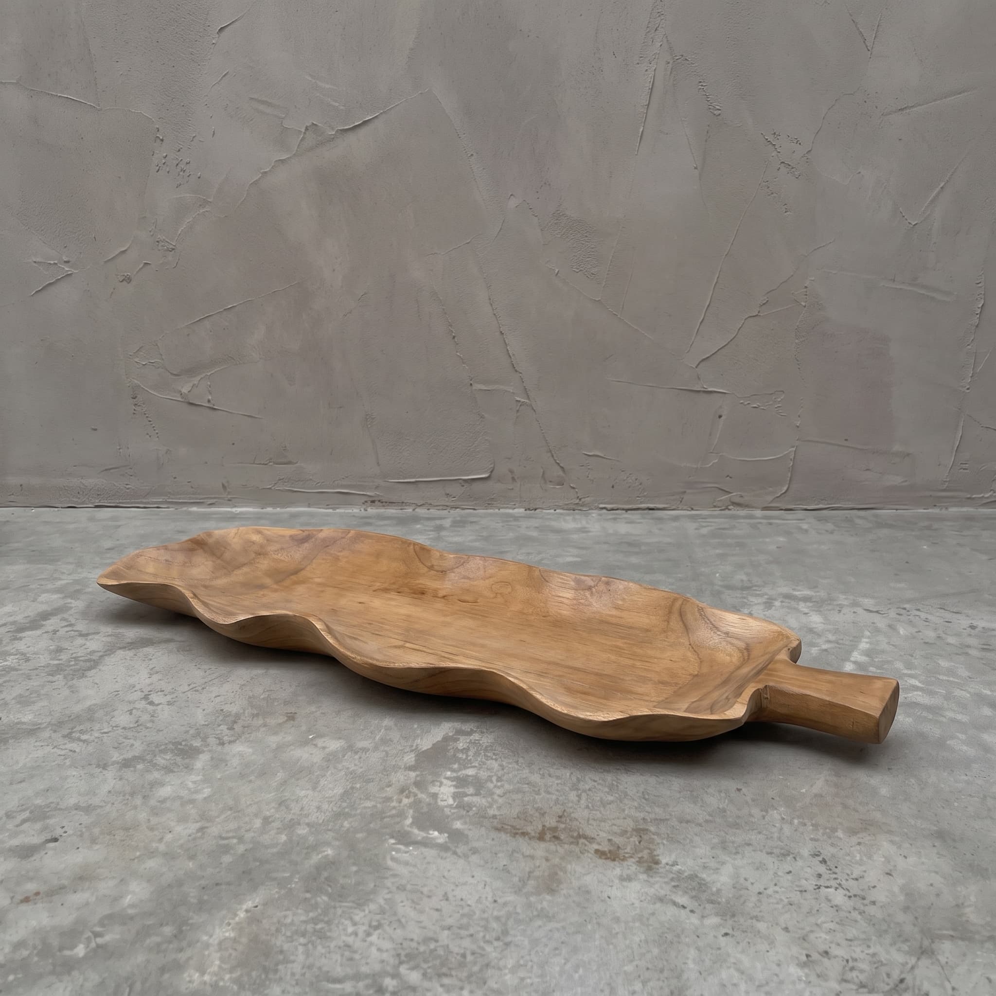 Bladvormige fruitschaal van gerecycled teak met golvende rand en een korte steel als handvat, op een betonvloer
