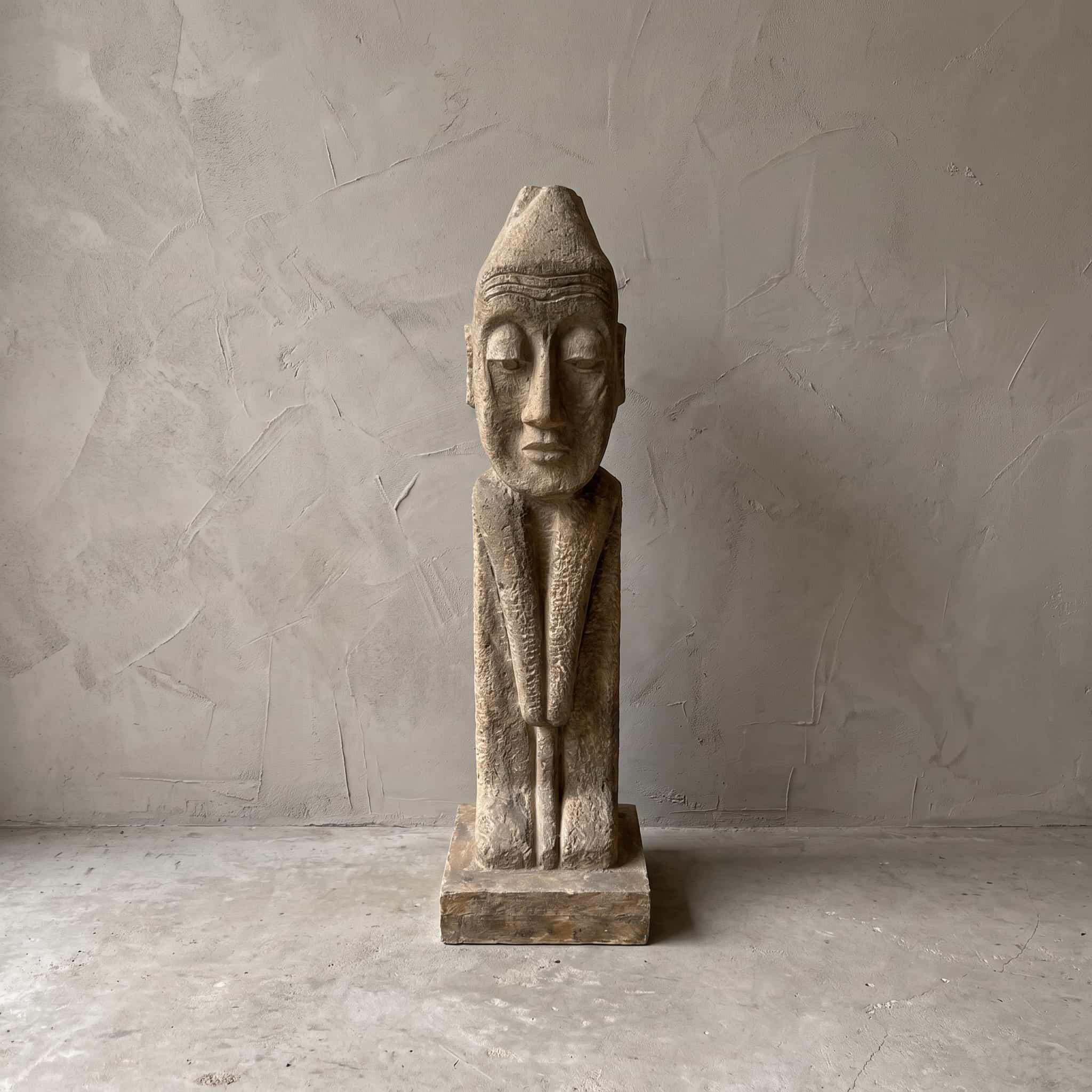 Beeld Konggo 1 van polystone, zandkleurig, vooraanzicht van de figuur met puntmuts op een lage voet voor een grijze wand