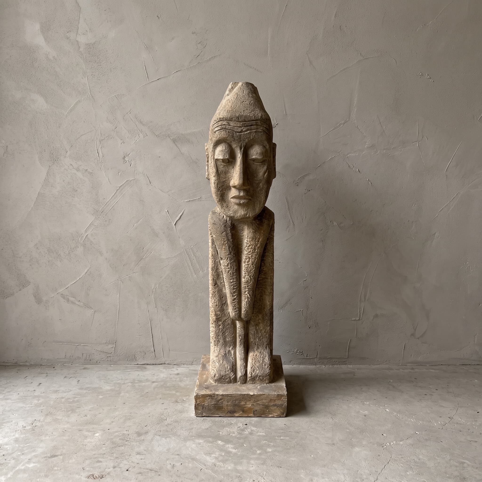 Beeld Konggo 1 van polystone, zandkleurig, vooraanzicht van de figuur met puntmuts op een lage voet voor een grijze wand