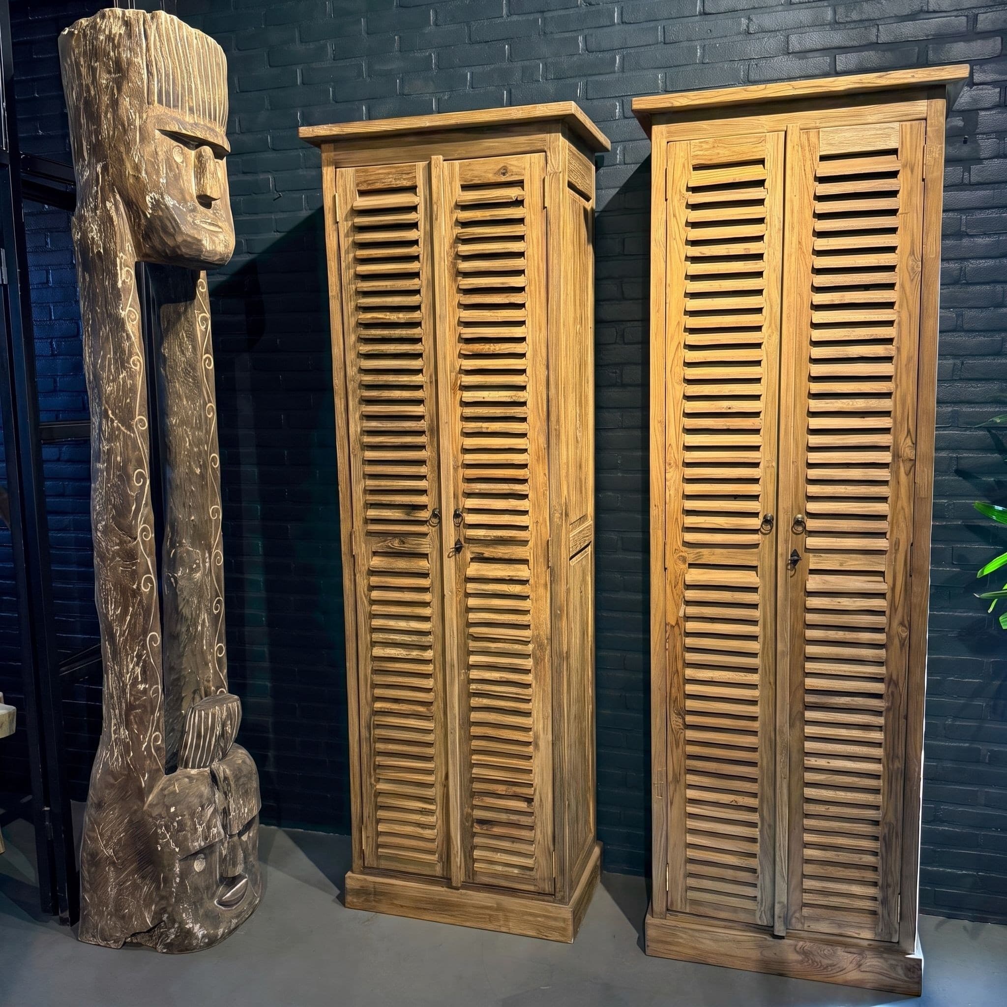 Kast Bali van gerecycled teak, 220 cm hoog, twee kasten met lamellendeuren tegen een donkere bakstenen muur, naast een houten beeld