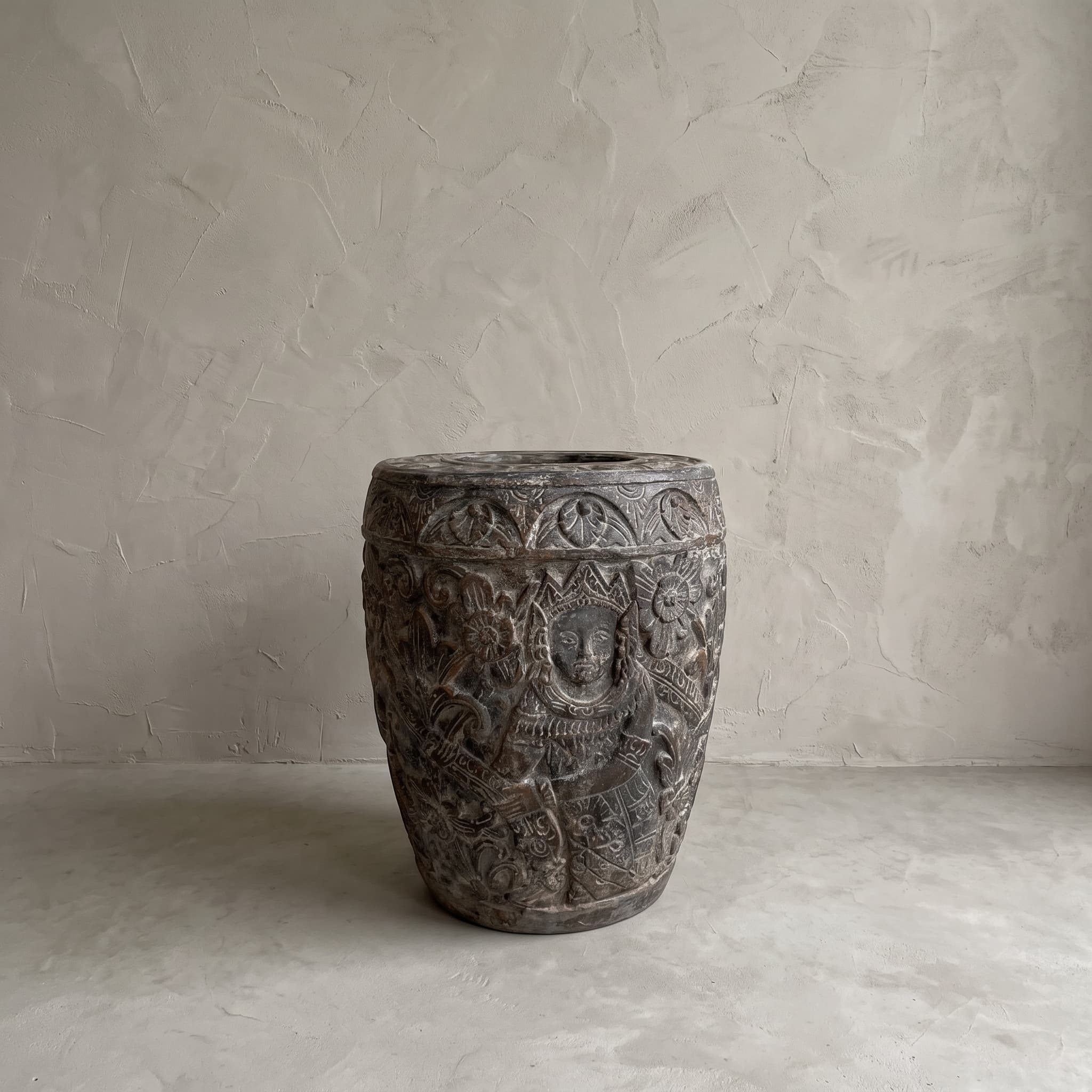 Pot Ukir van polystone, donker reliëf van een figuur met hoofdtooi tussen bloemen en bladeren, op een betonvloer