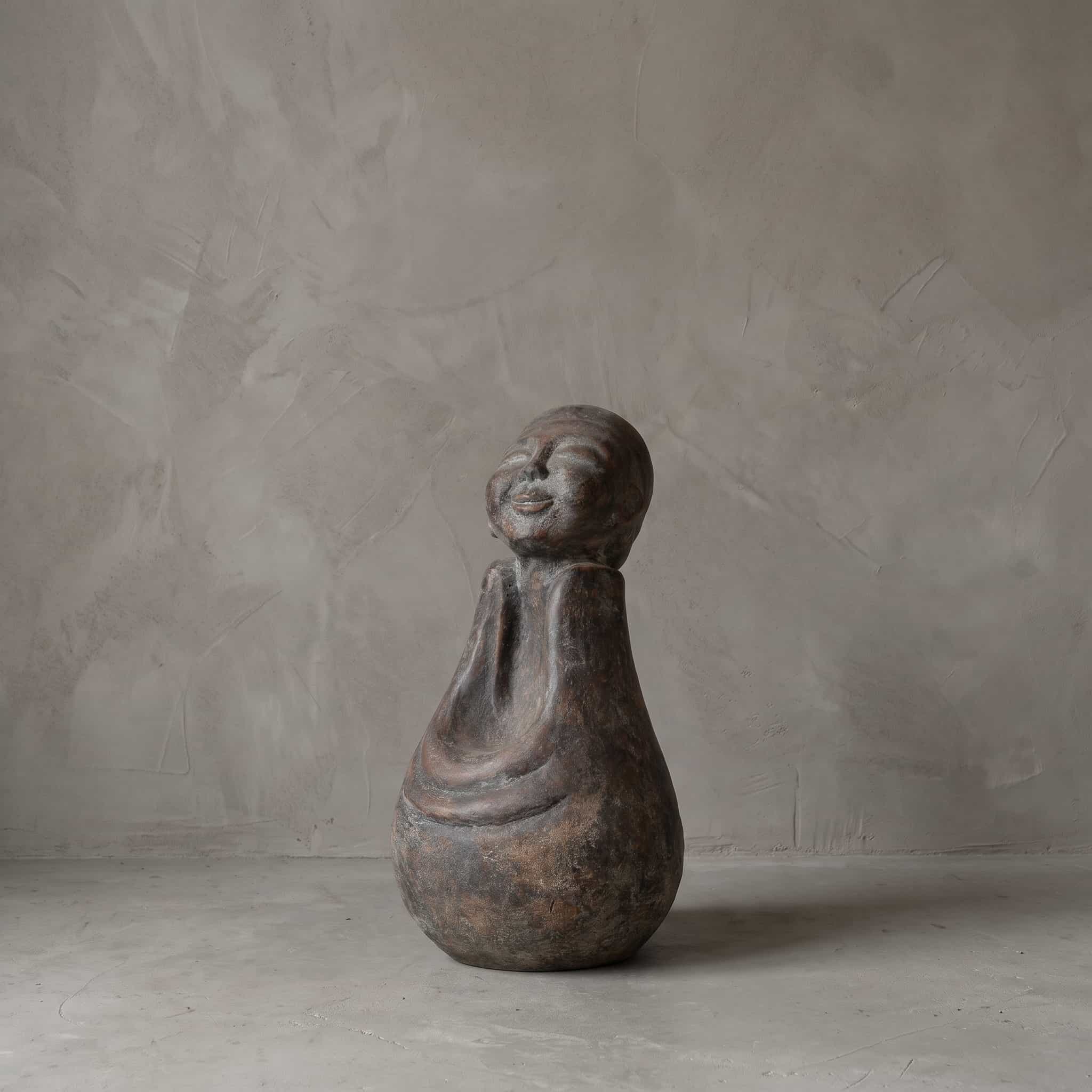 Boeddha Senyum van polystone, schuin vooraanzicht van de glimlachende figuur met gevouwen handen voor een grijze wand