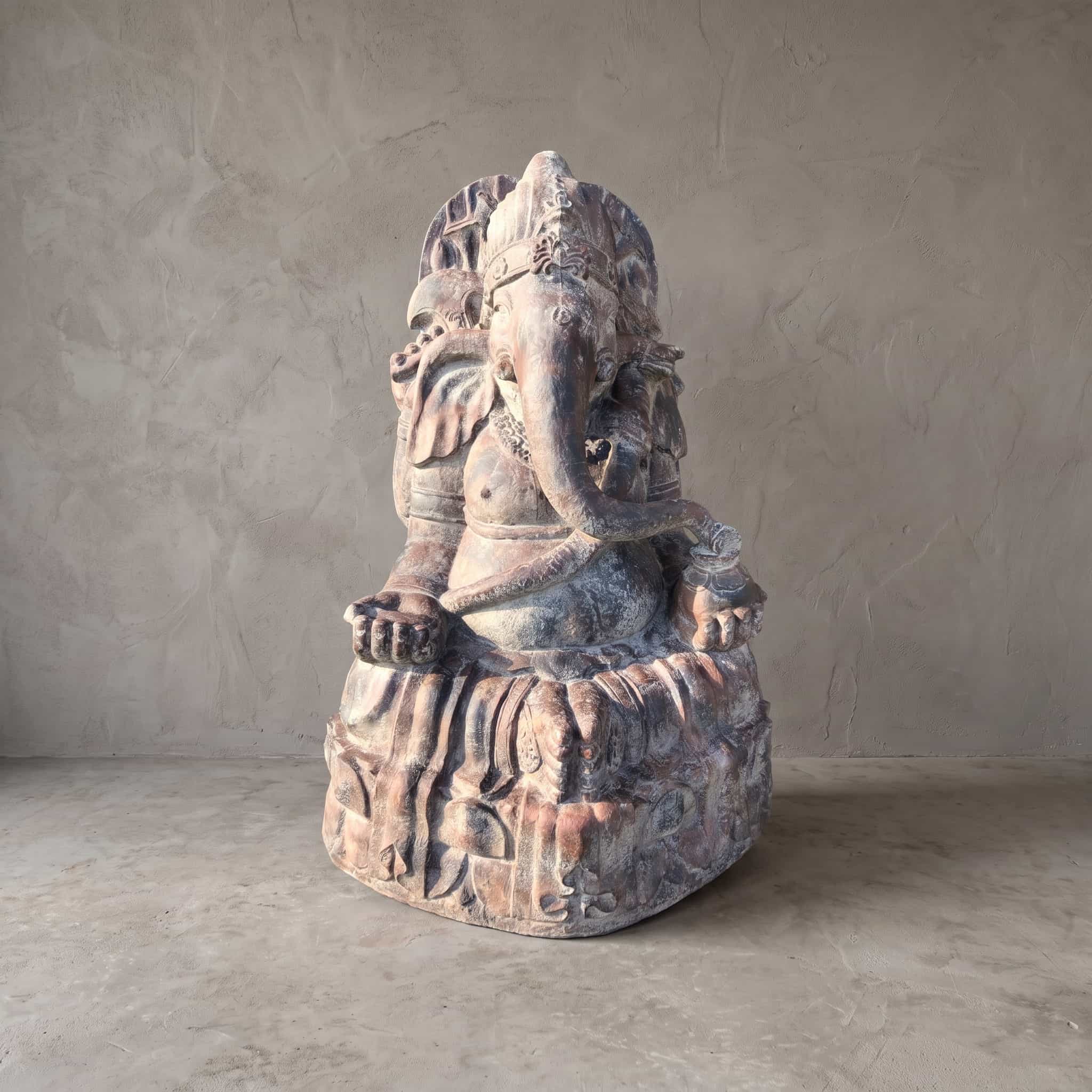 Beeld Ganesha van polystone, schuin vooraanzicht van de zittende Ganesha op een voet met reliëf voor een grijze wand