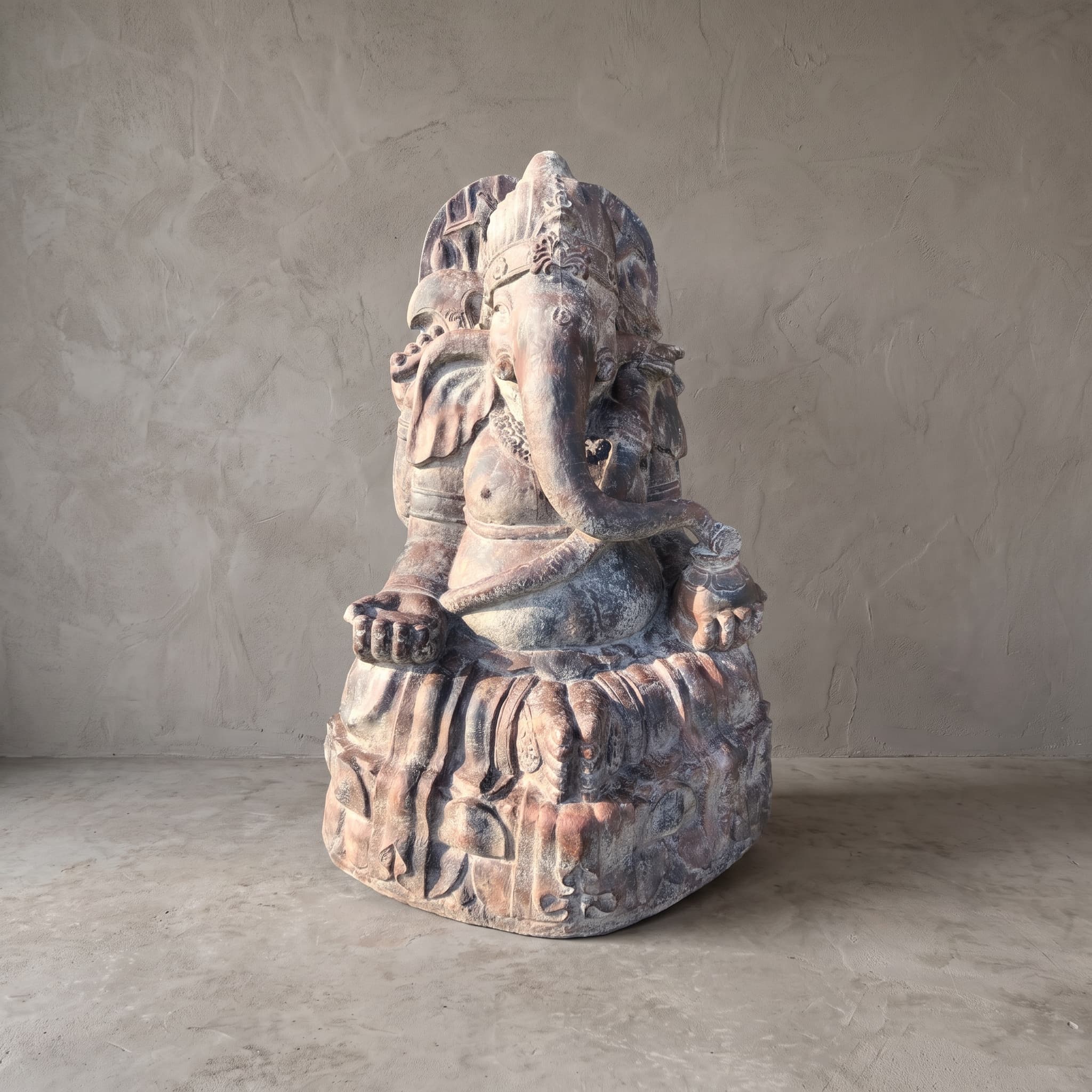 Beeld Ganesha van polystone, schuin vooraanzicht van de zittende Ganesha op een voet met reliëf voor een grijze wand