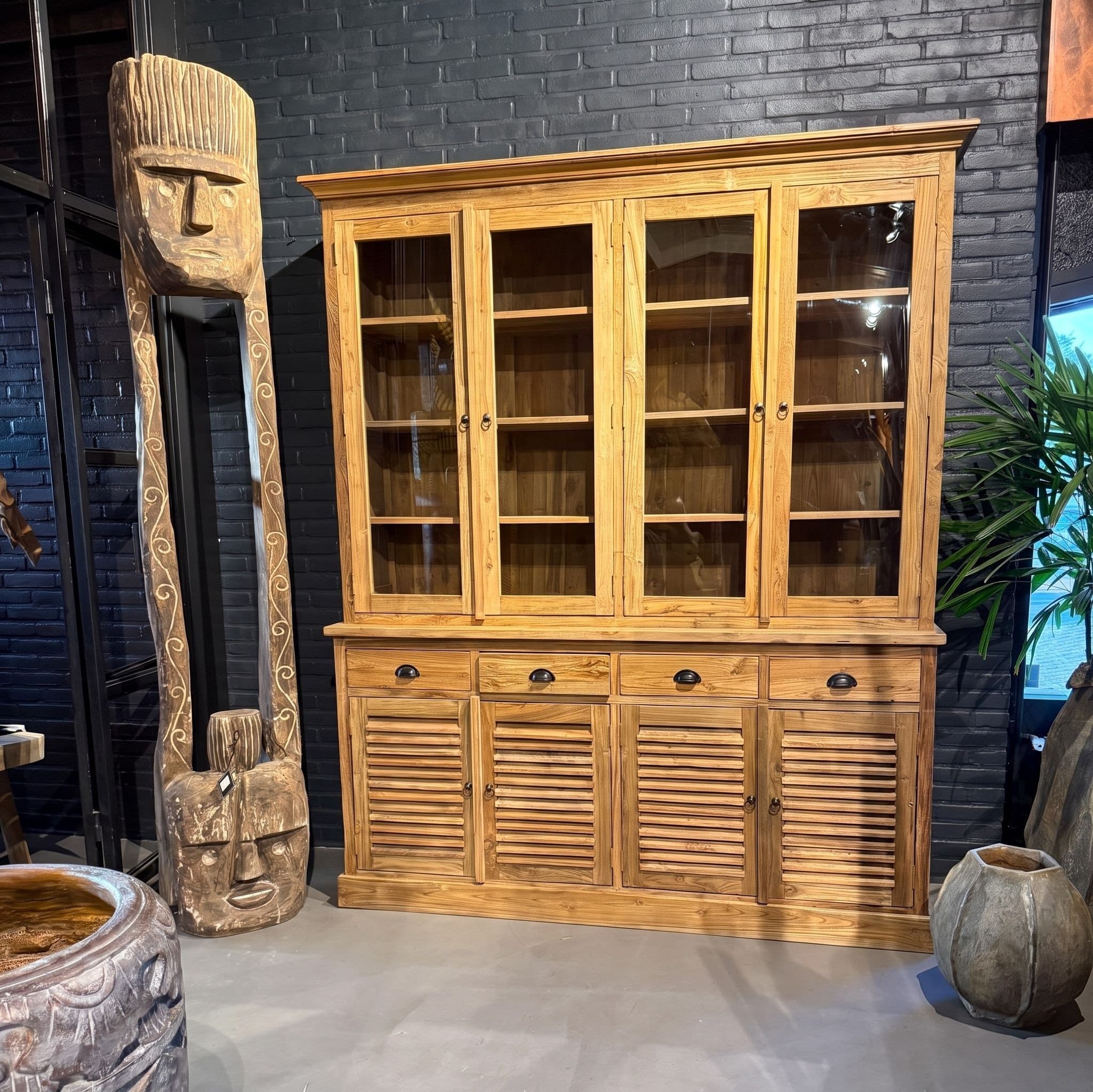 Kast Bali Tumpuk van gerecycled teak in de showroom, glazen deuren boven en lamellendeuren onder, tegen een donkere bakstenen muur