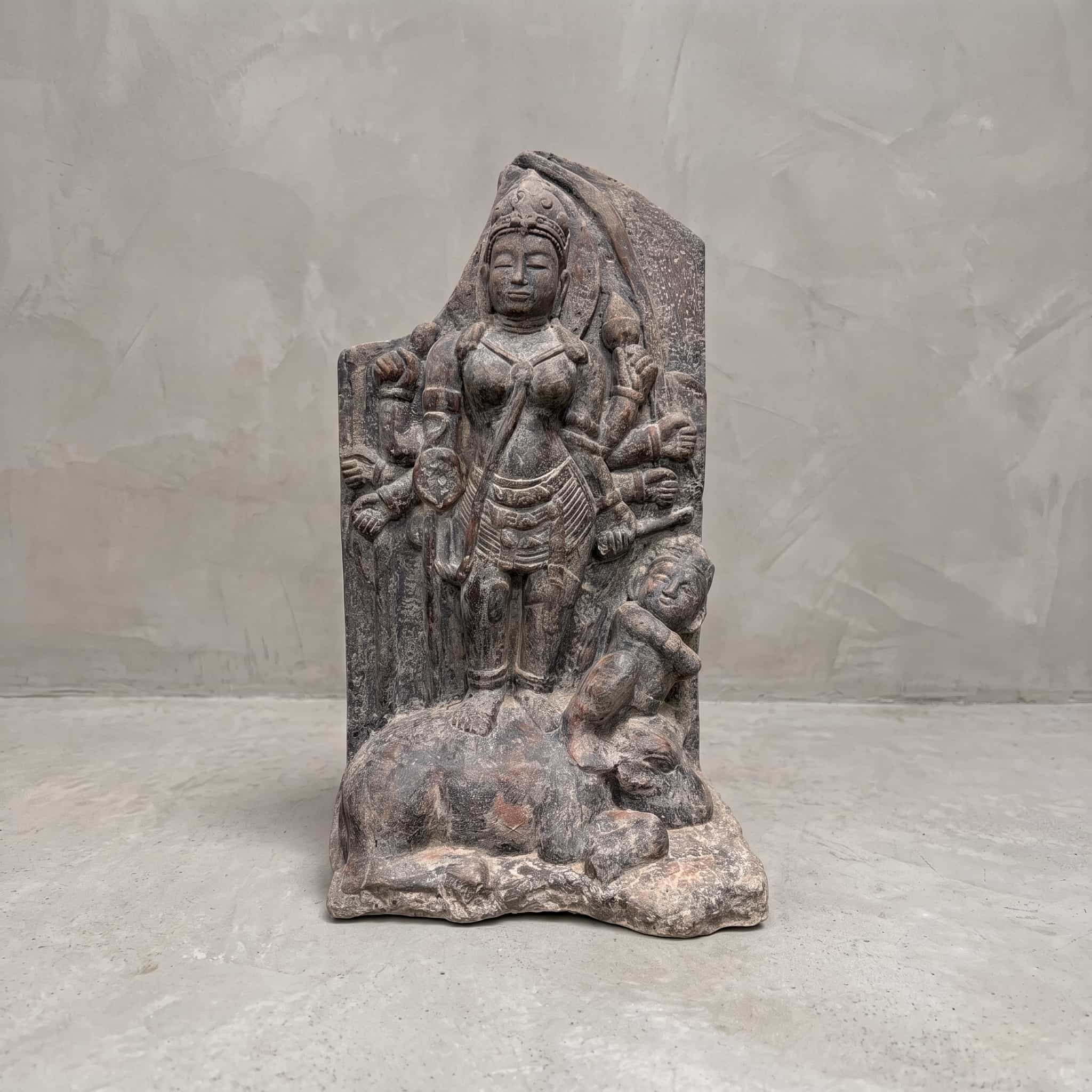 Beeld Tebeng van polystone, vooraanzicht van de figuur met meerdere armen op een liggend dier voor een grijze wand