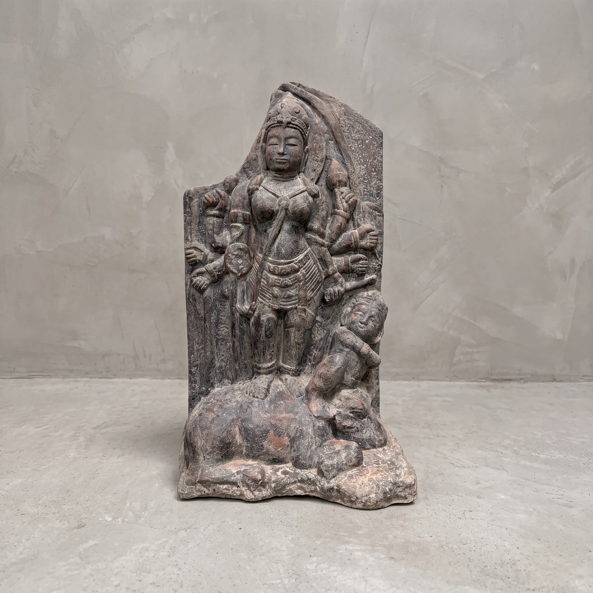 Beeld Tebeng van polystone, vooraanzicht van de figuur met meerdere armen op een liggend dier voor een grijze wand